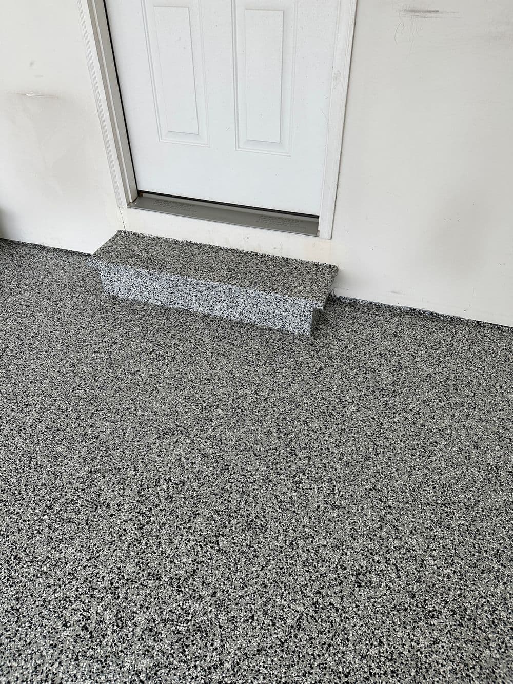 Granite-patterned floor with a gray step leading to a white door in a garage setting.