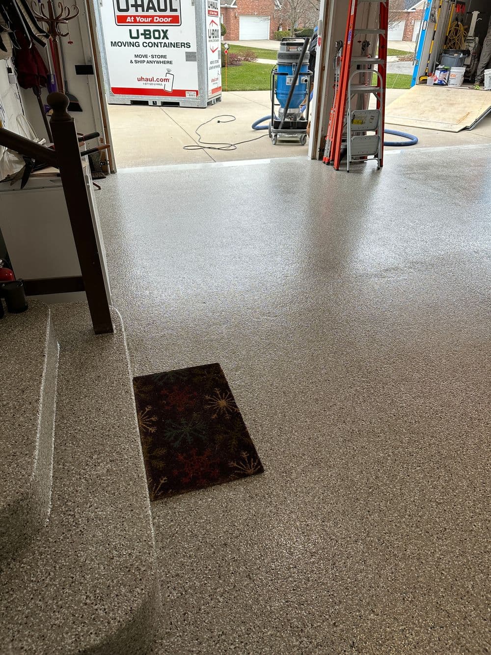 Garage interior with textured epoxy floor, mat at entrance, and moving containers in background.